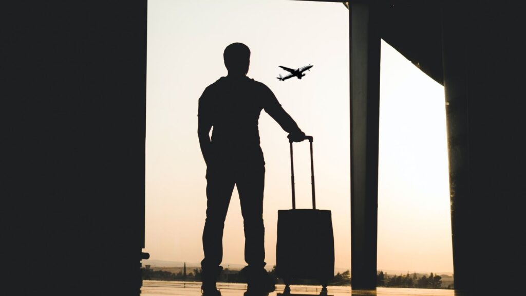 Silhouette of person at airport with suitcase looking out the window at a plane flying away