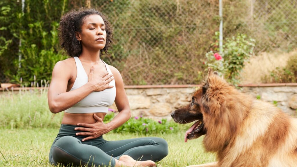 Black women sitting cross-legged outside with hand on heart and belly, doing yogic breathing