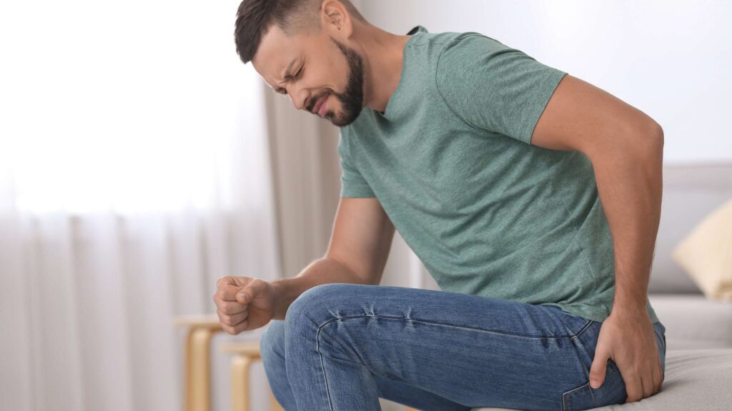 Man sitting on a couch grimacing in pain as he clunches his left hip/glute area.