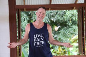 yogi aaron (a man) smiling with arms out in a gesture of receiving wearing a teacher that says live pain free